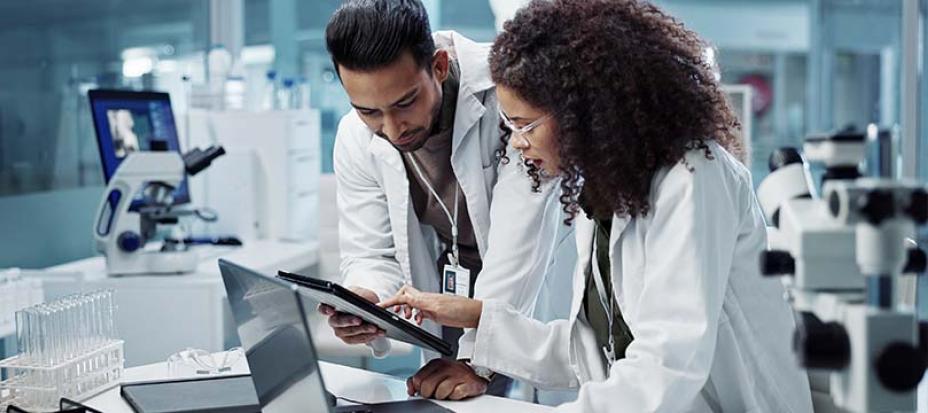 An image of two scientists in lab coats who are standing in a lab looking at a tablet.