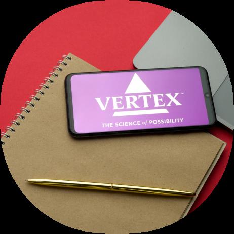 Vertex Pharmaceuticals logo visible on smartphone display sitting on top of notebook and laptop on desk