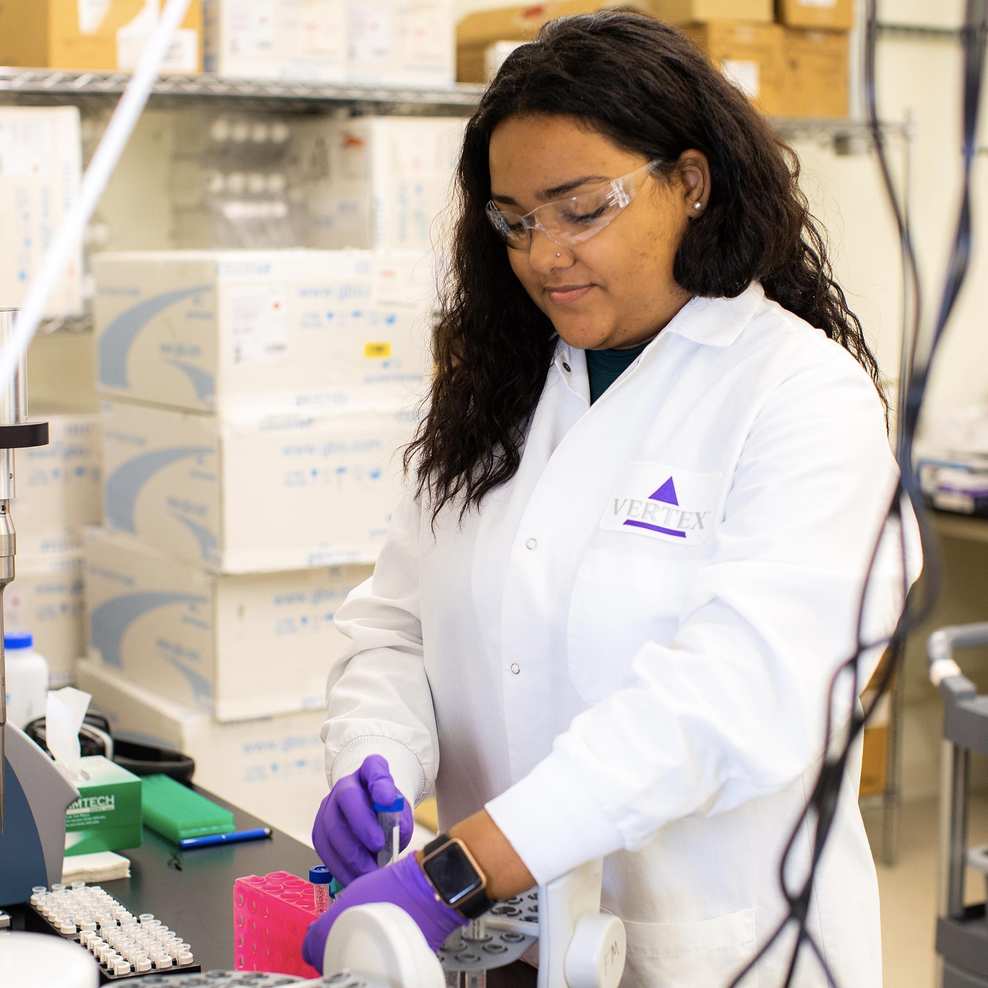 An image of a high school student wearing a white Vertex Pharmaceuticals lab coat while working with lab equipment to conduct an experiment