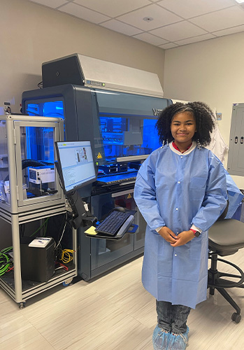 An image of Samara Seaton smiling while wearing blue scrubs standing in front of lab equipment in the San Diego Learning Lab.