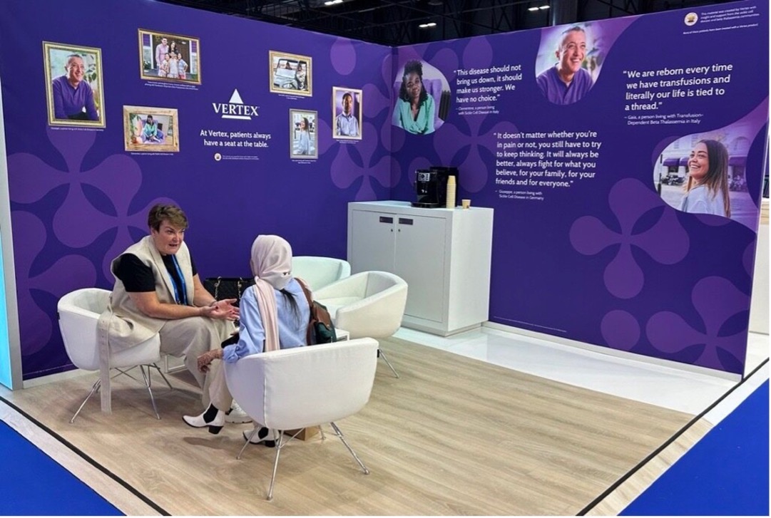 An image of the Vertex Pharmaceuticals booth at the EHA conference in 2024. Two people are sitting in white chairs having a conversation in front of purple walls that feature quotes from patients involved with Vertex.