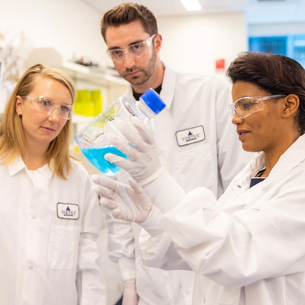 A Vertex scientist holds up a jar containing a blue liquid for 2 other employees to observe