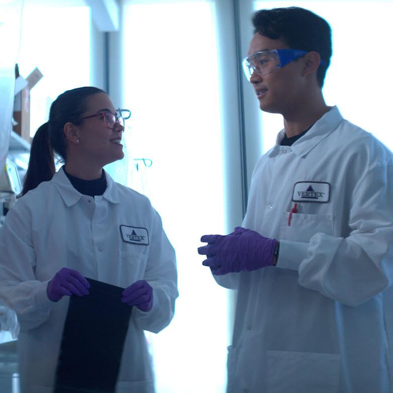 An image of two Vertex scientists wearing white lab coats conversing while surrounded by lab technology